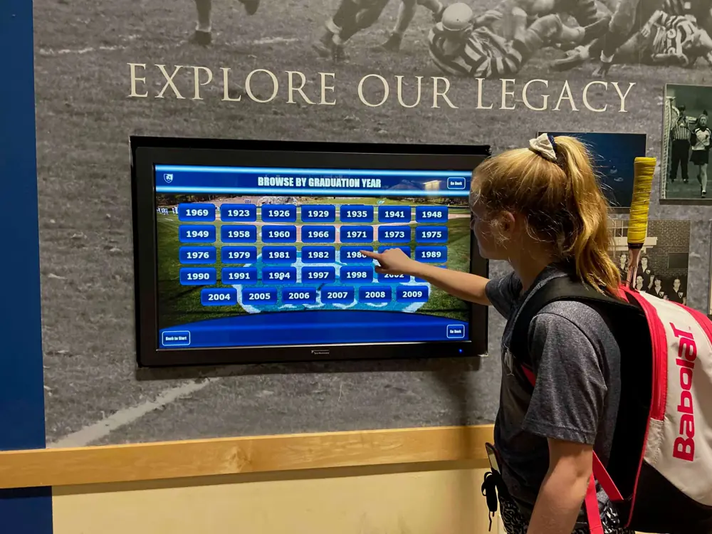 A student browses a touchscreen hall of fame by graduation year beneath the words Explore Our Legacy